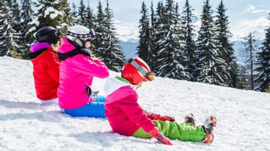 Bir grup insan kırmızı Kayak takım elbiseli bir dağın tepesinde kar üzerinde oturuyor. Bir aile kayakçı kar üzerinde oturmuş dinleniyor