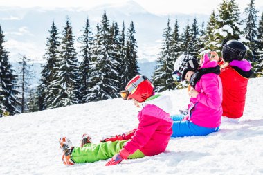 Bir grup insan kırmızı Kayak takım elbiseli bir dağın tepesinde kar üzerinde oturuyor. Bir aile kayakçı kar üzerinde oturmuş dinleniyor