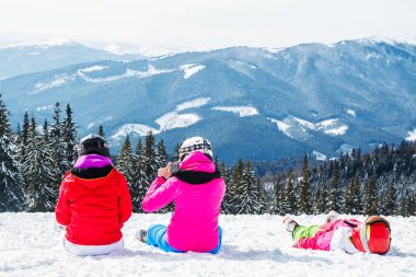Bir grup insan kırmızı Kayak takım elbiseli bir dağın tepesinde kar üzerinde oturuyor. Bir aile kayakçı kar üzerinde oturmuş dinleniyor