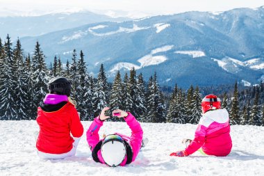 Bir grup insan kırmızı Kayak takım elbiseli bir dağın tepesinde kar üzerinde oturuyor. Bir aile kayakçı kar üzerinde oturmuş dinleniyor