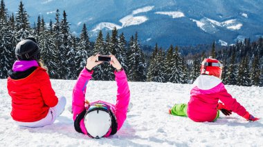 Bir grup insan kırmızı Kayak takım elbiseli bir dağın tepesinde kar üzerinde oturuyor. Bir aile kayakçı kar üzerinde oturmuş dinleniyor
