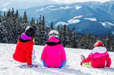 Bir grup insan kırmızı Kayak takım elbiseli bir dağın tepesinde kar üzerinde oturuyor. Bir aile kayakçı kar üzerinde oturmuş dinleniyor