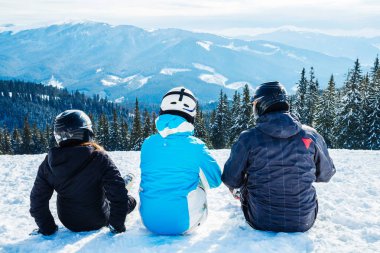 Üç kişi Kayak elbiseli dağ ve mesafe içine seyir tepesinde kar üzerinde oturuyor. Aile dağın dinlenme kayakçı. Arkadan görünüm