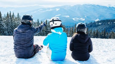 Üç kişi Kayak elbiseli dağ ve mesafe içine seyir tepesinde kar üzerinde oturuyor. Aile dağın dinlenme kayakçı. Arkadan görünüm