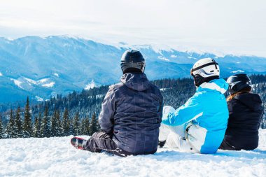 Üç kişi Kayak elbiseli dağ ve mesafe içine seyir tepesinde kar üzerinde oturuyor. Aile dağın dinlenme kayakçı. Arkadan görünüm