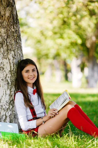 Молода школярка в шкільній формі сидить на траві під деревом з книгами — стокове фото