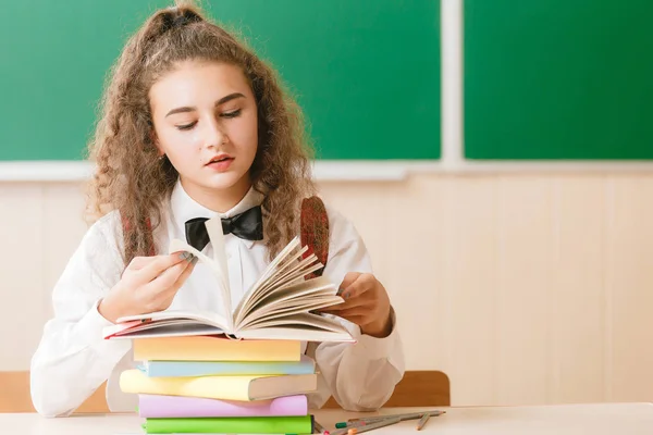 Écolière en uniforme scolaire assise à son bureau avec des livres et des crayons sur le fond d'un tableau vert — Photo