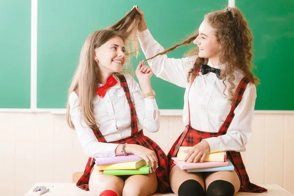 Two funny schoolgirls in school uniforms are sitting on the background of a green board on the desk with books.