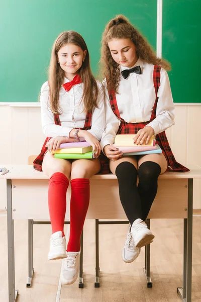 Two funny schoolgirls in school uniforms are sitting on the background of a green board on the desk with books.