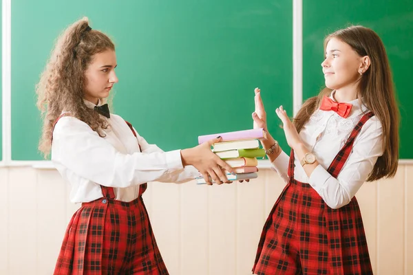 two funny schoolgirls in school uniform are standing with books on the background of the school board.