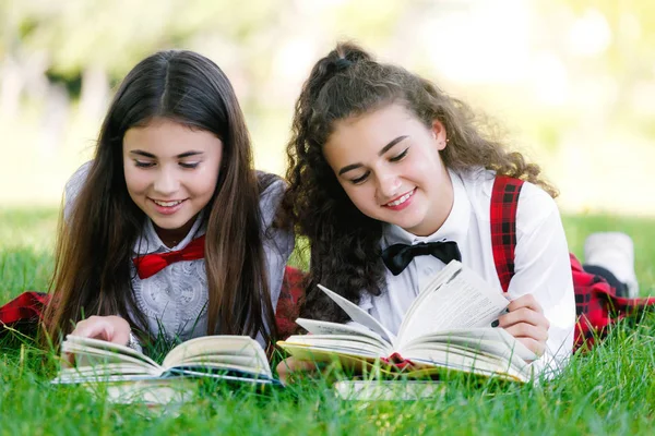 two funny schoolgirls are lying on the grass and reading books. Girls, girlfriends, sisters are taught lessons in nature.