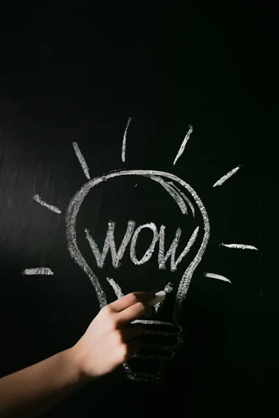 closeup female hand drawing a white chalk light bulb on a black board with the word wow.