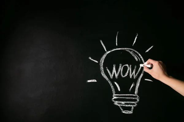 closeup female hand drawing a white chalk light bulb on a black board with the word wow.