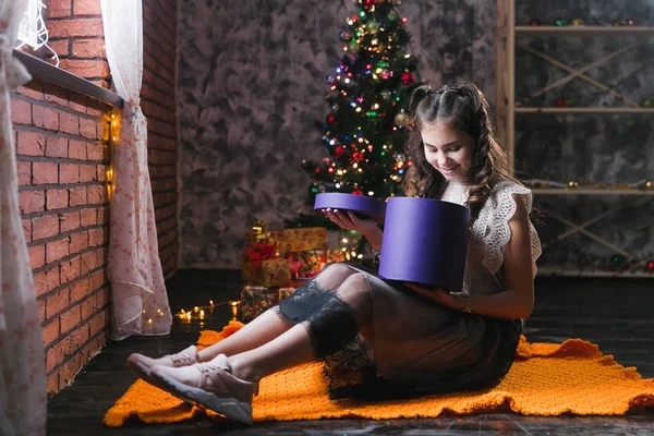 little girl in a dress sits on the floor at home near the Christmas tree and holds a box with gifts.brunette girl sits in a dark room and opens a lilac box with a gift