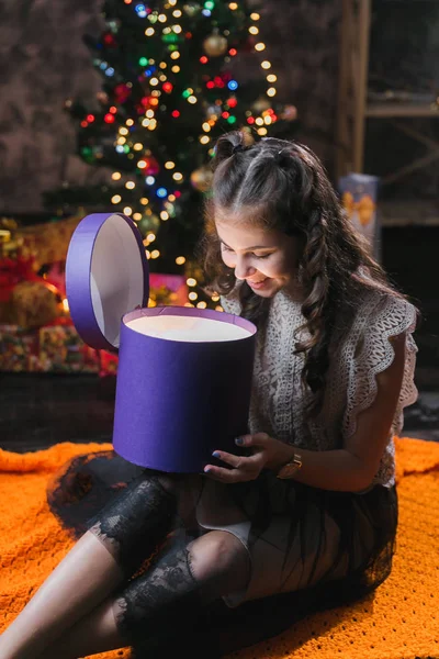 little girl in a dress sits on the floor at home near the Christmas tree and holds a box with gifts.brunette girl sits in a dark room and opens a lilac box with a gift