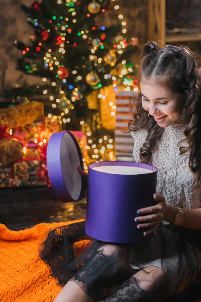 little girl in a dress sits on the floor at home near the Christmas tree and holds a box with gifts.brunette girl sits in a dark room and opens a lilac box with a gift