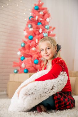 Küçük sarışın kız bir kırmızı kazak ve damalı rahim karşı pembe bir Noel ağacı zemin katta parlak bir odada oturuyor ve büyük bir beyaz yumuşak yastık hugs ve kameraya gülümseyen süre görünüyor. Noel Tema