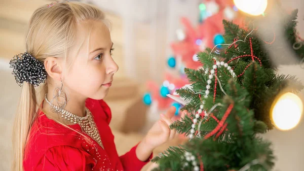 クリスマス ツリーの近くに赤いセーターの金髪少女が立っています 子供がおもちゃでクリスマス ツリーを飾る — ストック写真