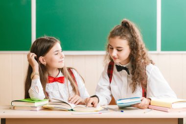 iki brun.ette kız öğrenciler tahtanın arka planda kitaplar ile masasında sınıfta oturup.kızlar bir okul dersi oturup