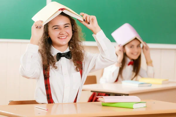 two schoolgirls are sitting at the desk in the classroom at the lesson. A girl sits at a table with books and learns.schoolgirl covered her head with a book sitting at the table in the classroom