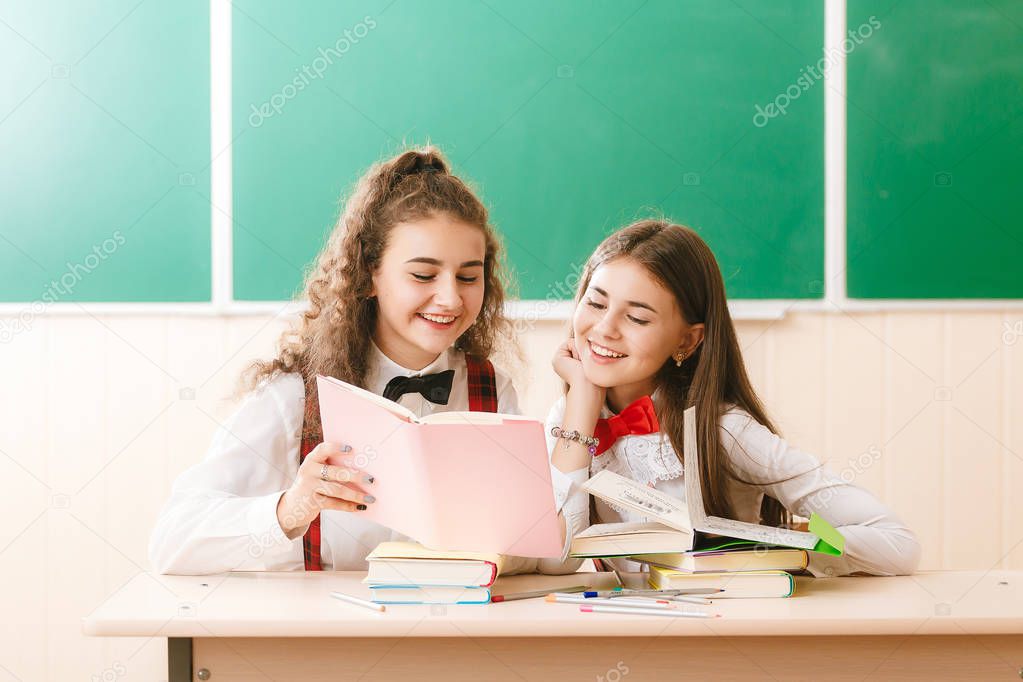 two brun.ette schoolgirls sit in the classroom at her desk with books on the background of the blackboard.girls sit on a school lesson