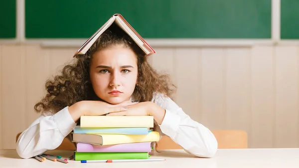 Blackboard Young 女子高生黒板の背景に彼女の机に座って 彼女の頭に本を持っての背景に関する書籍を学校の机に座ってかわいいブルネットの女子高生 — ストック写真