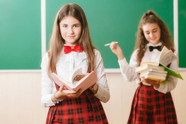 Duas alunas bonitas em uniforme escolar ficar com livros em sala de aula contra o fundo do quadro — Fotografia de Stock