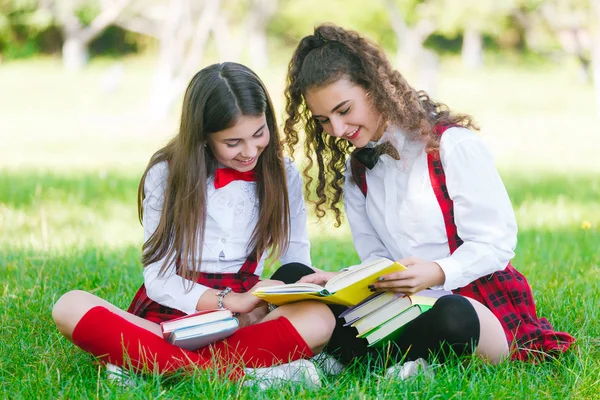 two schoolgirls in school uniforms sit with books in the park. Schoolgirls or students are taught lessons in nature