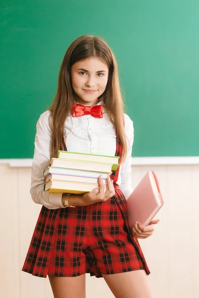 Alumno con uniforme escolar de pie en una clase escolar frente a la pizarra y sosteniendo libros de texto — Foto de Stock