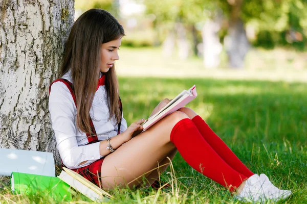 Студент у шкільній формі викладає уроки під відкритим небом. дівчина сидить на траві в парку і читає книгу . — стокове фото