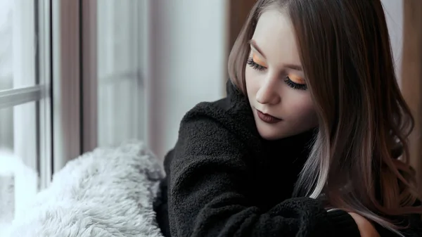 Ein schönes junges Mädchen sitzt mit geschlossenen Augen allein auf der Fensterbank. eine Frau im Pullover sitzt am Fenster — Stockfoto