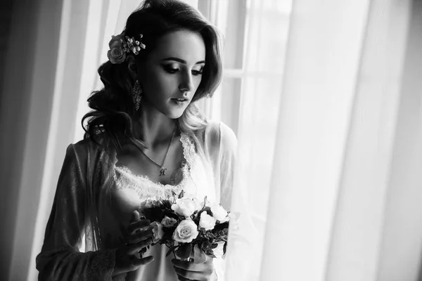 Bella ragazza con i capelli biondi in una stanza luminosa vicino alla finestra. Sposa in vestaglia con un bouquet — Foto Stock