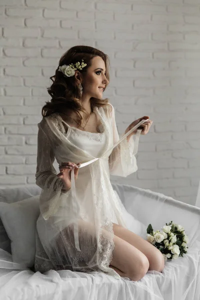 Bella ragazza con i capelli biondi in una stanza luminosa vicino alla finestra. Sposa in vestaglia con un bouquet — Foto Stock