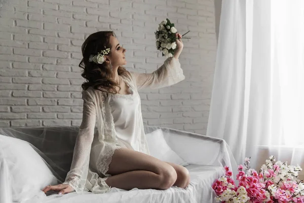 Mooi meisje met blond haar in een lichte kamer in de buurt van het raam. Bruid in dressing Gown met een boeket — Stockfoto