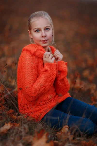 穿着橙色毛衣的金发女孩坐在秋天的森林或公园里 看着摄像机 — 图库照片
