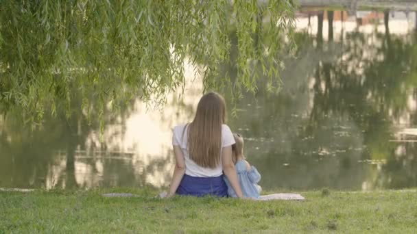 夕日の光の中で湖の近くの木の下に座ってお母さんと小さな女の子 木と夕日水に反映 幸せな家族の概念 — ストック動画