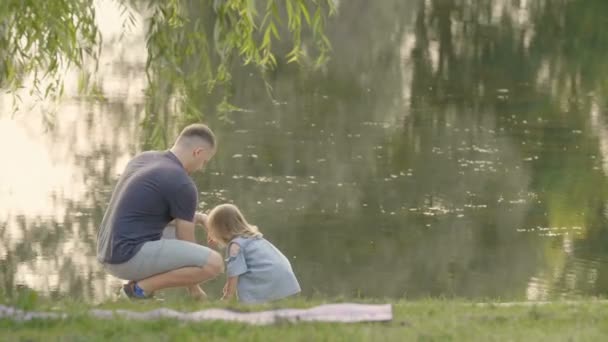 夕焼けの光の中で湖とフィード魚の近くの木の下に座っている父と娘 赤ちゃんの感情 幸せに笑います 木と夕日水に反映 幸せな家族の概念 — ストック動画