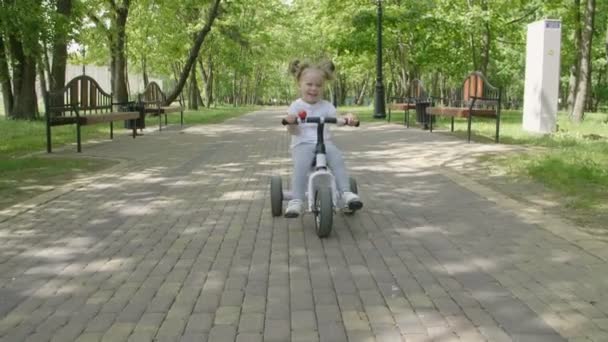 女婴在公园里骑自行车 快乐的孩子笑和笑 一个小女孩在城市公园骑自行车 慢动作 — 图库视频影像