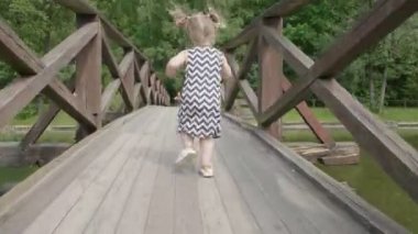 Küçük bir bebek kız ahşap bir köprü boyunca çalışan ve gün batımı ışığında annenin kollarına acele. Geri görünüm.