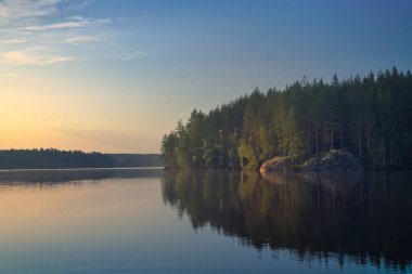 Yumuşak gün batımı ışığı sakin gölü yansıtır. Orman, kayalar ve gökyüzü barışçıl ve uyumlu bir manzara yaratmak için birleşir..