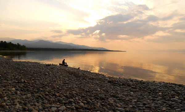 Człowiek siedzi sama na dziennik o ponad brzeg morza, Świt ciszy na jeziorze. — Zdjęcie stockowe