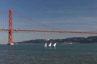 Portekiz'in Lizbon şehrinde arka planda 25 Nisan köprüsü ile Tagus Nehri'nde üç küçük yelkenli tekne