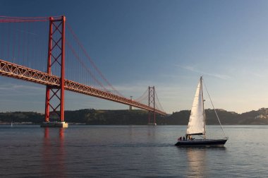 Portekiz'in Lizbon şehrinde Tagus Nehri üzerinde 25 Nisan Köprüsü'nün (Ponte 25 de Abril) yanından geçen yelkenli tekne; Portekiz'de seyahat konsepti
