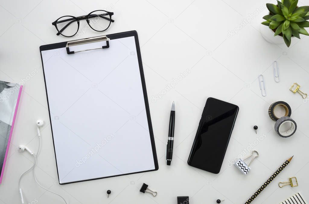 Flat lay, top view Home office desk workspace with office accessories including clipboard with blank paper space for lettering, black smartphone, glasses, paper clips, pen, pencil, pins, tape and