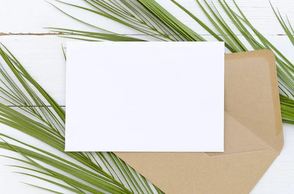 Minimal composition white blank card and envelope on palm leaves on a white wooden background. Mockup with envelope and blank card. Flat lay. Top view.