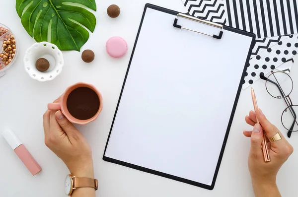 Flat lay clipboard mockup. Mãos femininas segura caneta dourada e xícara de café em branco folha de papel branco zombar. Escritório em casa com óculos, folhas verdes e doces . — Fotografia de Stock