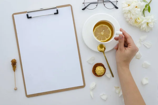Top view desk clip board mockup. womans hands holds cup of tea with lemon,workspace is decorated with white roses,crystals sugar, glasses and sweets. Morning concept. Flat lay mock up