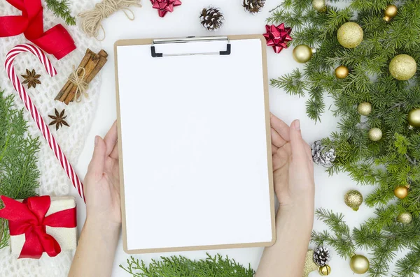 Mockup Christmas composition. pine cones fir branches, gift box, golden balls, candy on white background. Womens hands holds clipboard with blank paper sheet. Flat lay, top view notes to do list.