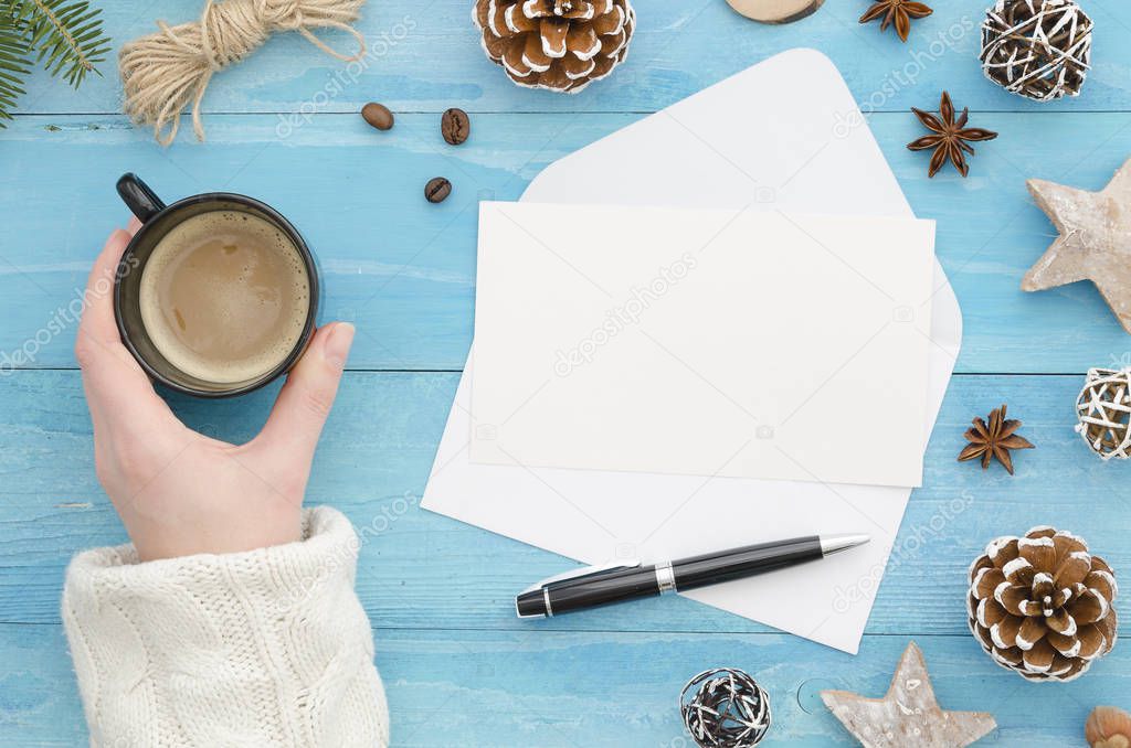 Mock up envelope. Female hand holding a cup of coffee on holiday workplace with Christmas gifts, New Year decorations, pine cone. Greeting card copy space top view