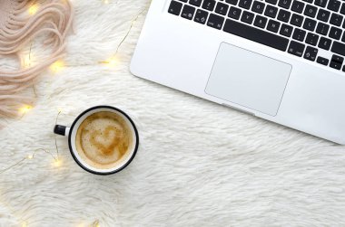 Sıcak sıcak kış çalışma alanıyla kahve ve dizüstü kürk kabarık beyaz zemin üzerine ışıklar garland ve eşarp. Üstten görünüm mockup